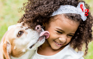Young child with curly hair smiling while hugging a small dog outdoors on grass.