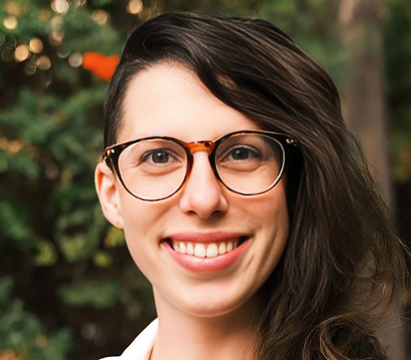 Smiling woman with dark hair and glasses in front of green foliage background.