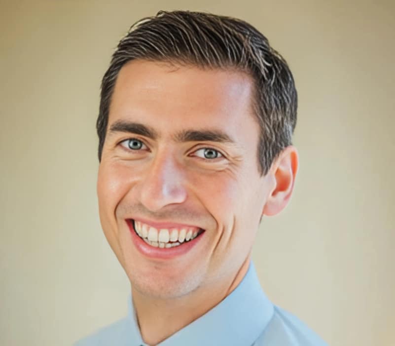 Professional headshot of smiling male dentist in light blue shirt against neutral background.